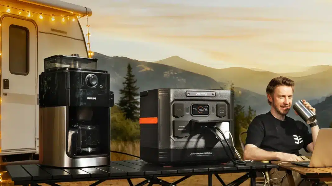 Jackery Explorer 1500 Ultra on a camp table beside a caravan, powering a coffee maker, while a man sits at a laptop with a drink