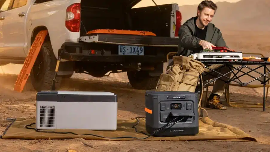 Jackery Explorer 1500 Ultra on a ground mat next to a portable fridge, with a man cooking on a camp table beside a ute's open tailgate