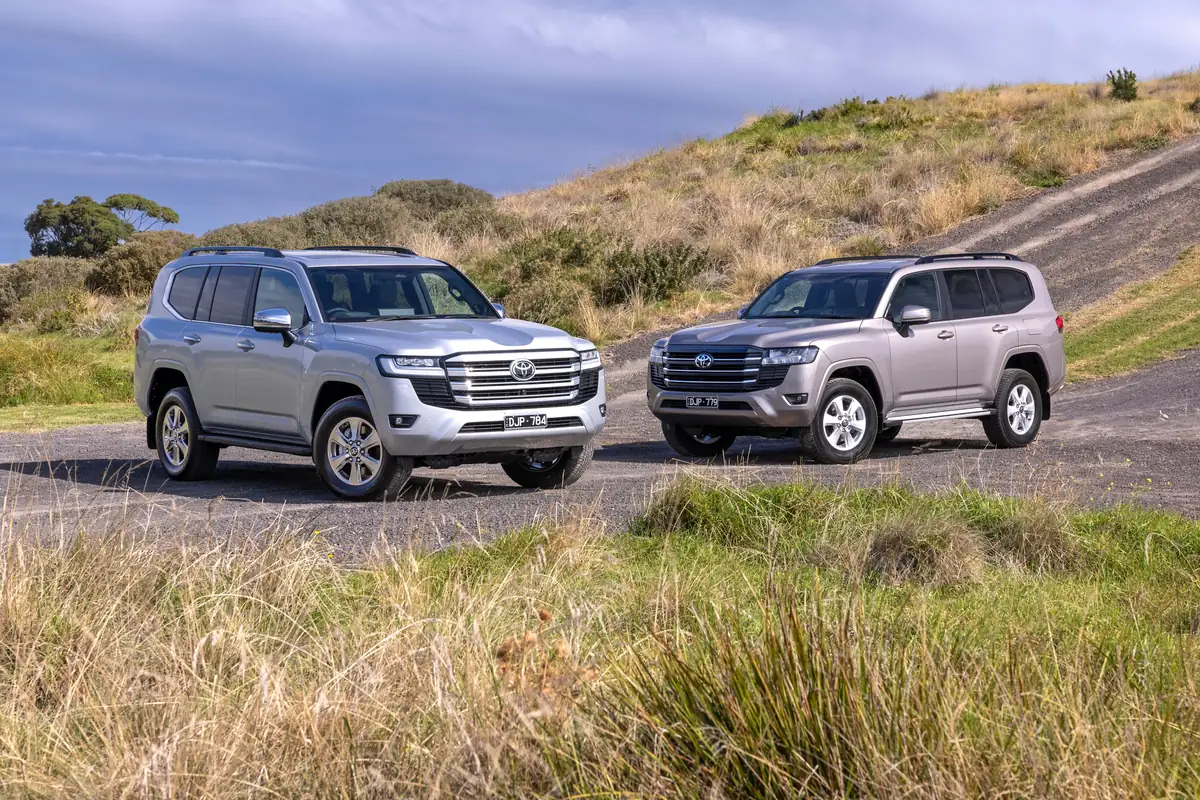 Two Toyota LandCruiser 300 Series wagons, one silver and one bronze, parked on a gravel road among grassy hills
