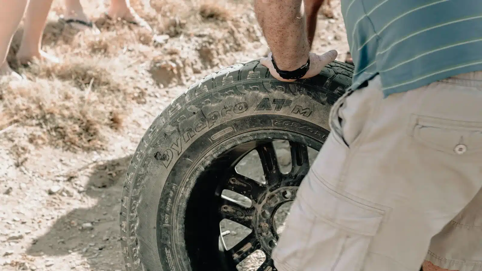 Man in shorts holding an all-terrain tyre on its alloy wheel on dry, dusty ground