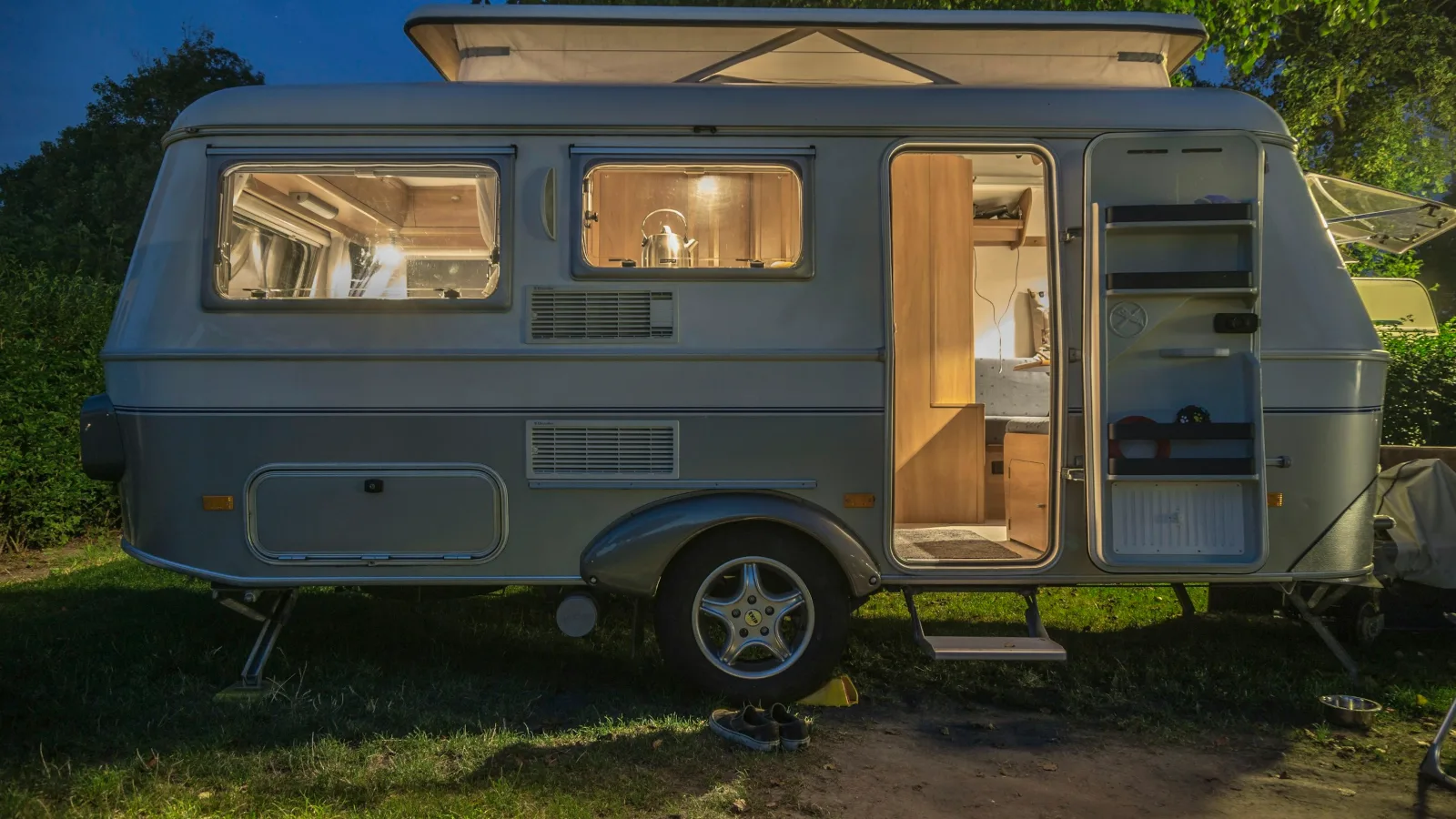 Silver older-style caravan with its door open and interior lights on, parked on grass at dusk