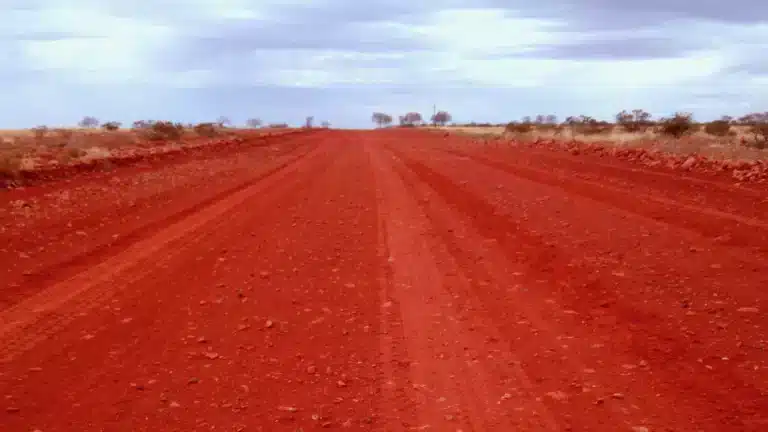 Straight red gravel road with wheel ruts running through low scrub on the Oodnadatta Track in South Australia