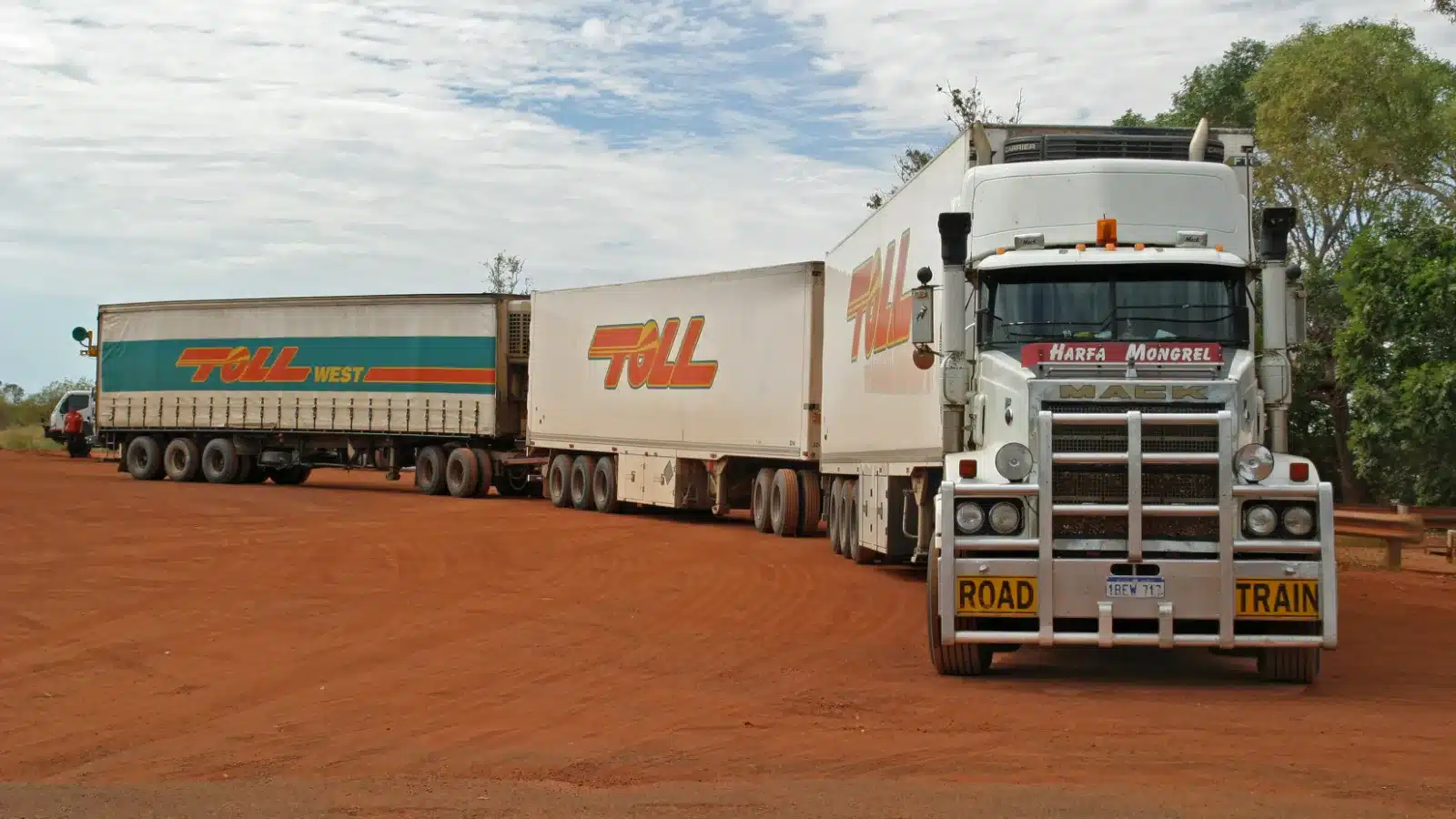 Triple road train with a white prime mover parked on red gravel beside trees in Western Australia