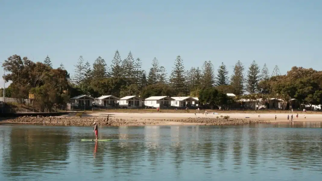 Row of white cabins and Norfolk Island pines behind a sandy beach, with a paddleboarder on calm water